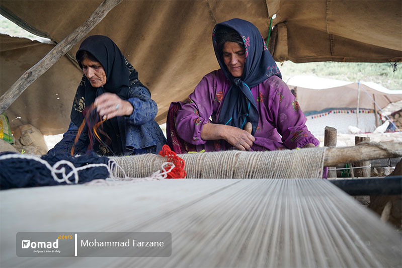 two nomad women are weaving a Persian carpet while sitting under their tent
